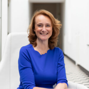 Een glimlachende vrouw in een blauwe jurk, zittend in een witte stoel in een gang met zwart-wit geblokte vloeren en witte muren.