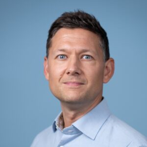 Een man met kort haar, gekleed in een lichtblauw shirt tegen een blauwe achtergrond.