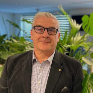 Man van middelbare leeftijd met een bril glimlachend in een kantooromgeving, gekleed in een grijze blazer en een geruit overhemd, met kamerplanten op de achtergrond.