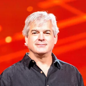 Een man van middelbare leeftijd met grijs haar staat zelfverzekerd op het podium, gekleed in een zwart shirt met een kleurrijke onscherpe achtergrond.