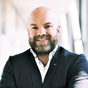 Een bebaarde man met een kaal hoofd in een donker pak en wit overhemd staat glimlachend met zijn armen over elkaar in een helder verlichte gang.