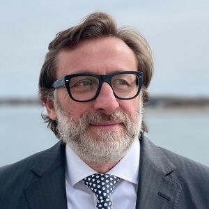 Buiten staat een man met baard en bril in pak en stropdas met stippen, met de zee op de achtergrond.