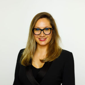 Een professionele vrouw met een bril en een zwarte blazer glimlacht zelfverzekerd tegen een witte achtergrond.