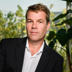 Een man van middelbare leeftijd in een zwart pak en een wit overhemd, zittend met een serieuze uitdrukking, voor wazig groen gebladerte.