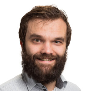 Een man met een baard en een lichte glimlach draagt een lichtgekleurd overhemd met een patroon en een donkere kraag, poserend tegen een witte achtergrond.