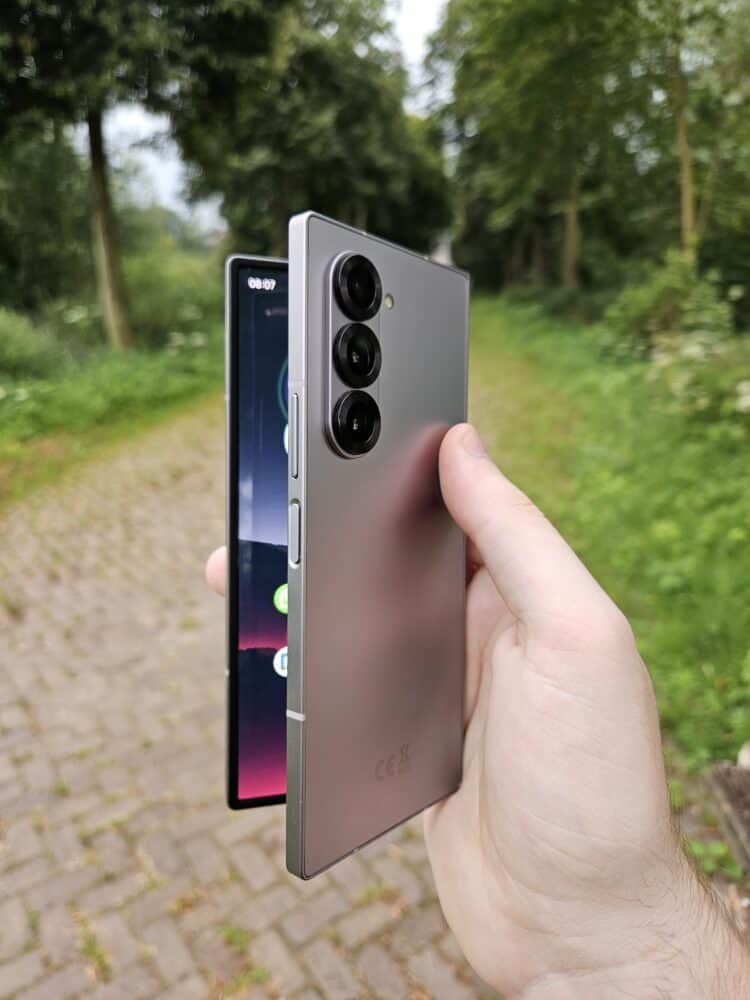 Een persoon houdt een opvouwbare smartphone vast met drie camera's op de achterkant, gedeeltelijk uitgevouwen, in een buitenomgeving met een pad en groen.