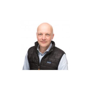 Een man met een kaal hoofd, gekleed in een lichtblauw shirt en een zwart gewatteerd vest, staat met zijn gezicht naar de camera gericht tegen een effen witte achtergrond.