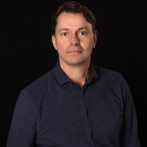 Een man met kort donker haar en een donker overhemd staat tegen een zwarte achtergrond en kijkt recht in de camera.