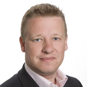 Een man met kort blond haar en blauwe ogen, gekleed in een zwart krijtstreeppak en een lichtroze overhemd, kijkt recht in de camera tegen een effen witte achtergrond.