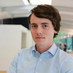 Een man met donkerbruin haar en een lichtblauw overhemd staat binnen, met op de achtergrond een wazig kantoor.
