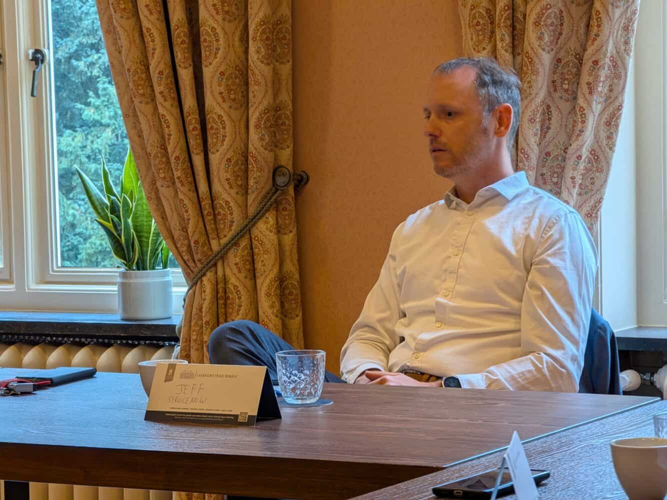 Een persoon in een wit shirt zit aan een tafel met een glas en een naambordje. De kamer heeft gordijnen met patronen en een potplant bij het raam.