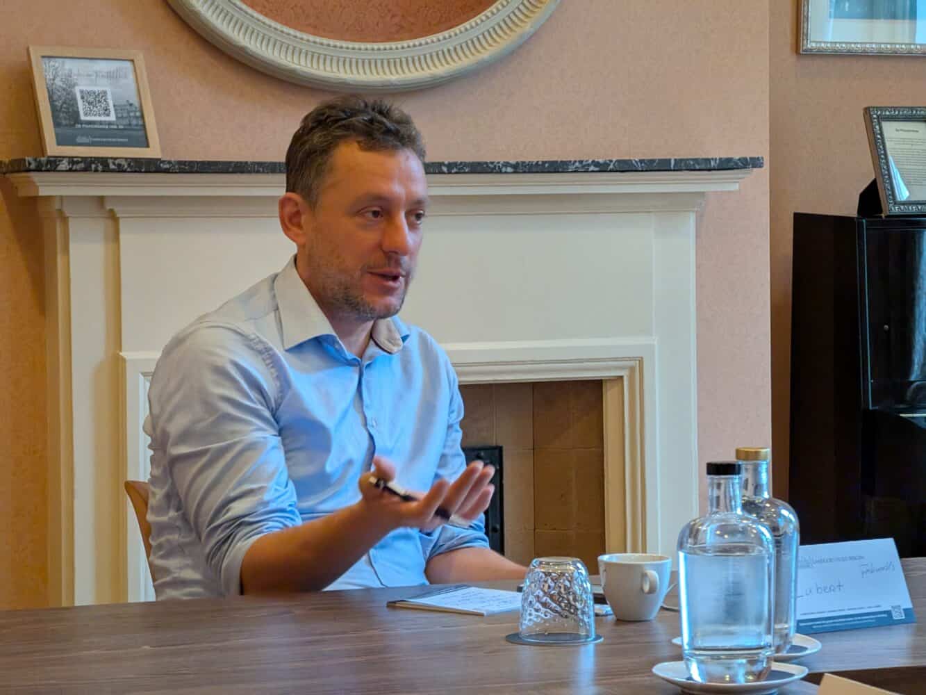 Een man in een lichtblauw shirt zit aan een tafel en gebaart met zijn rechterhand. Er staan een kopje, een glas en een fles op tafel. Een open haard en ingelijste items staan op de achtergrond.