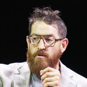 Een man met een bril en een baard, die een microfoon vasthoudt, zit op een podium met een rood-zwarte achtergrond.