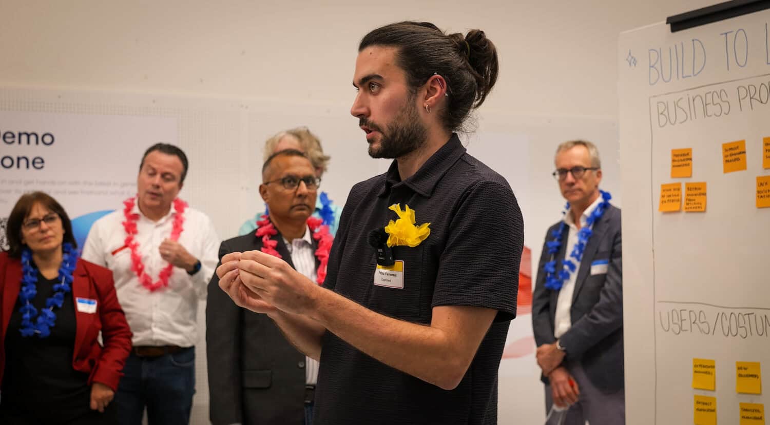 Een bewuste persoon spreekt een klein publiek toe in een kamer, waar de aanwezigen kleurrijke leis dragen en aandachtig luisteren. Rechts staat een whiteboard met plaknotities waarop brainstormideeën worden vastgelegd.