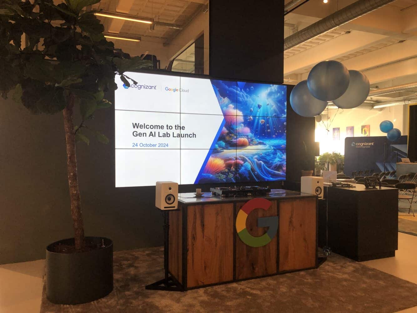 Het Gen AI Lab Launch-evenement bestaat uit een zaal met grote schermen waarop details van het evenement worden getoond, een houten podium versierd met de logo's van Google en Cognizant en decoratieve ballonnen.