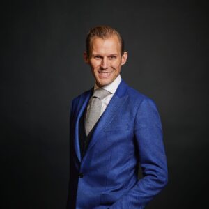 Een persoon in een blauw jasje en stropdas staat tegen een donkere achtergrond, lacht en poseert met één hand in zijn zak.