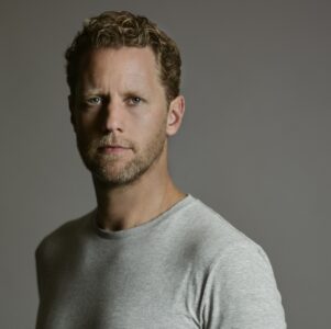Een man met krullend haar en een baard draagt een grijs T-shirt en staat tegen een effen achtergrond.