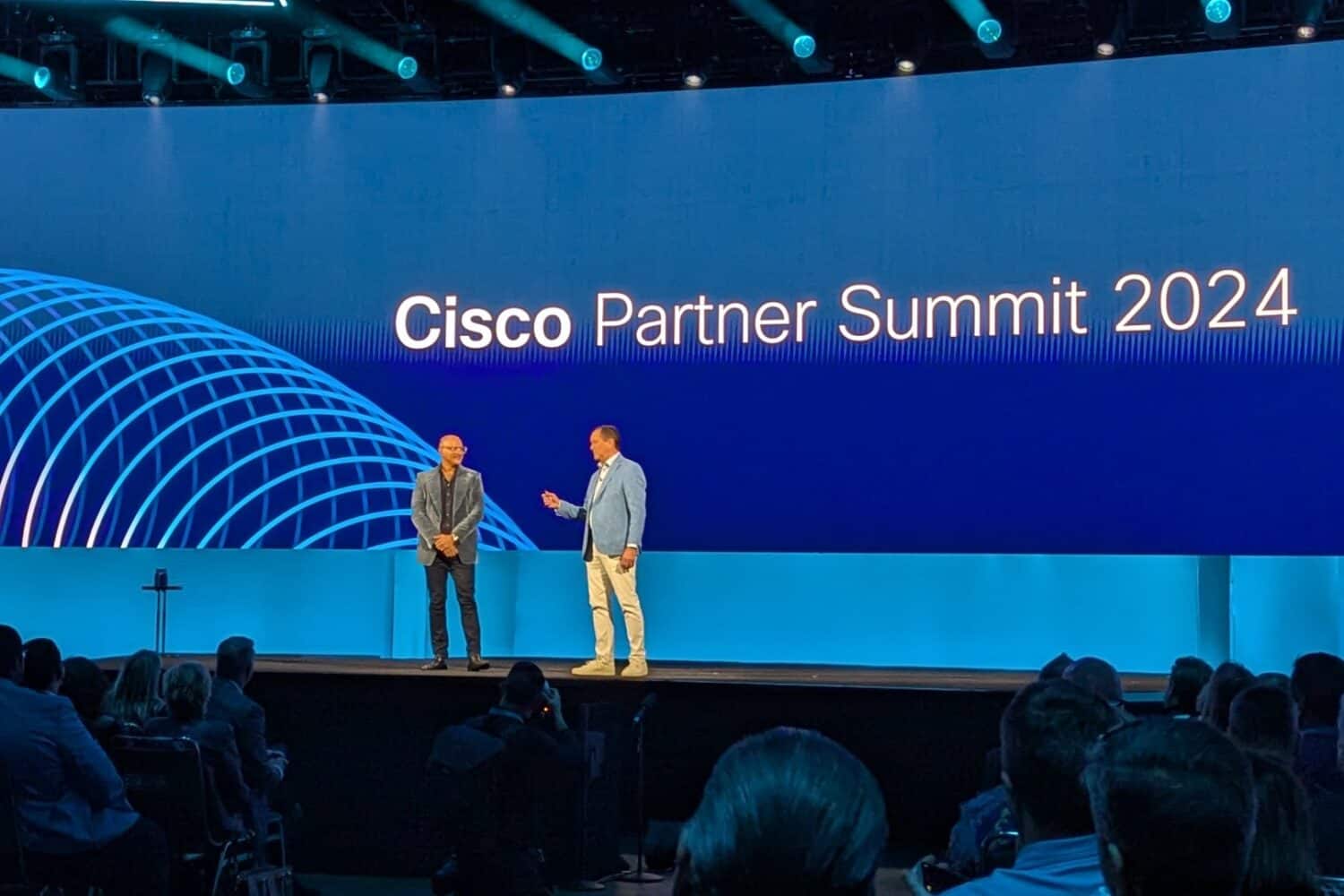 Twee sprekers op het podium van Cisco Partner Summit 2024, met een groot scherm waarop de naam van het evenement op de achtergrond wordt weergegeven. Publieksleden zitten op de voorgrond.