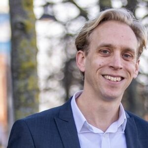 Een persoon met licht haar draagt een pak en lacht naar de camera, tegen een onscherpe achtergrond van bomen.