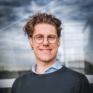 Een persoon met krullend haar en een bril lacht voor een reflecterende glazen achtergrond onder een bewolkte lucht.
