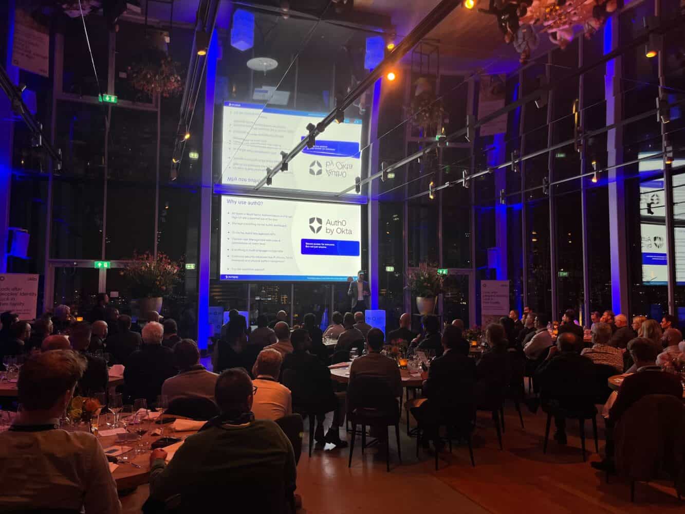 Mensen zitten in een schemerige ruimte en kijken naar een Okta-presentatie op grote schermen.