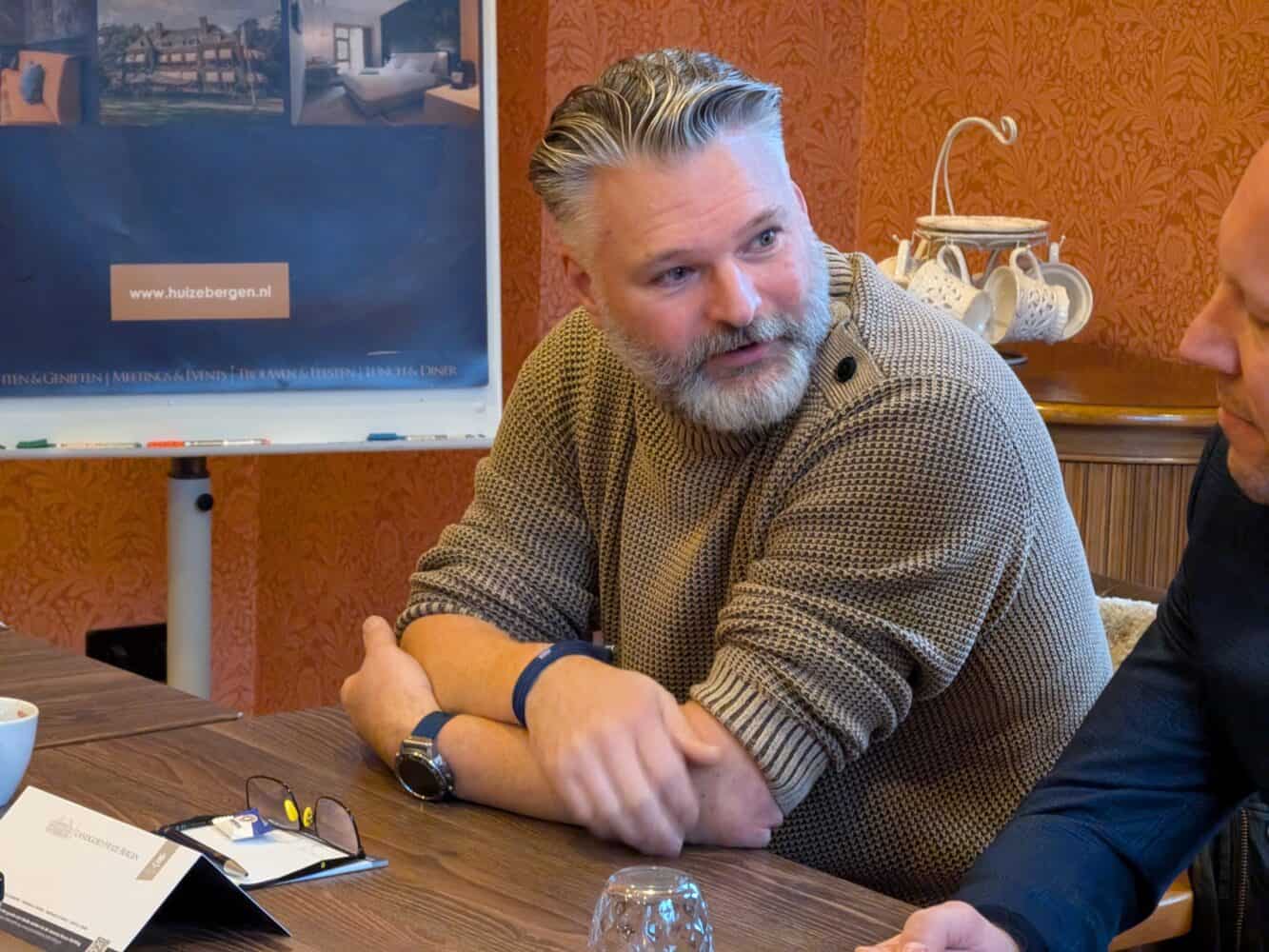 Een man met een baard zit aan een tafel en is in gesprek. Achter hem hangt een poster van een datacenter.