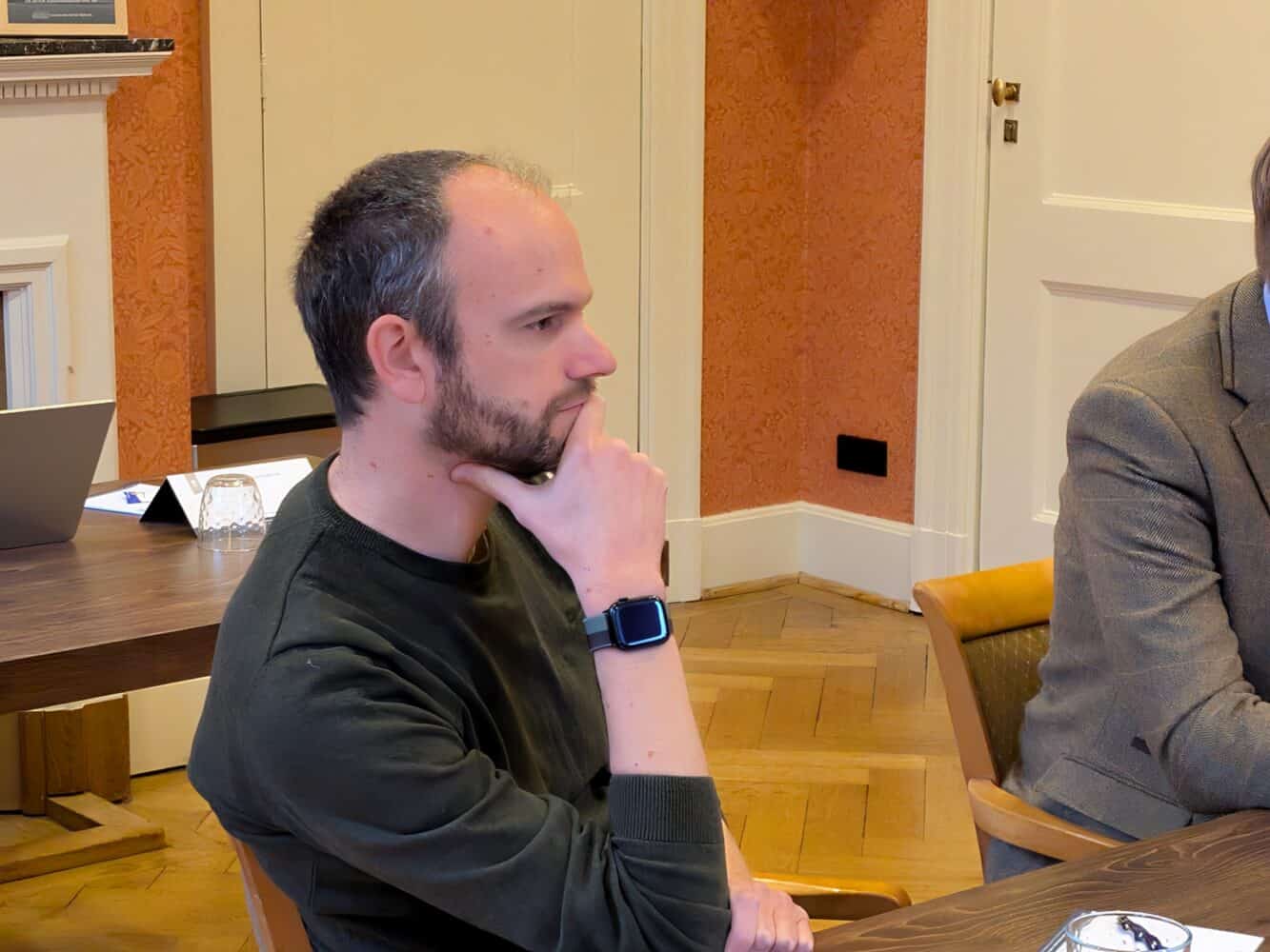 Een man met een baard, die een smartwatch draagt, zit aan een tafel en laat zijn kin op zijn hand rusten. De kamer, die doet denken aan een strak datacenter met zijn houten vloeren en levendige oranje muren, vormt een intrigerende achtergrond.