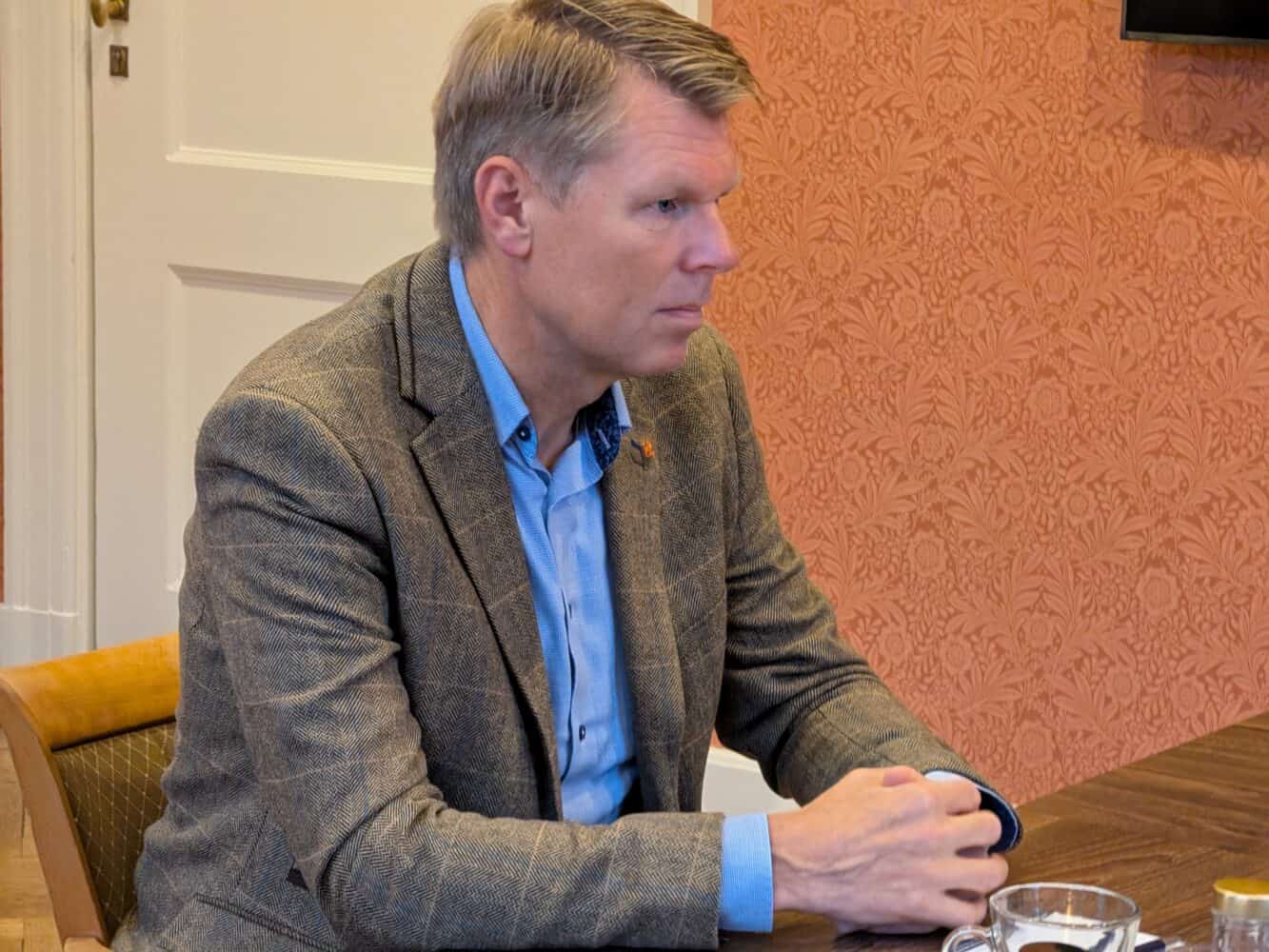 Een man in een geruit jasje en een blauw overhemd zit aan een houten tafel. De sfeer is als in een datacenter, in een kamer met een oranje muur met patroontjes.