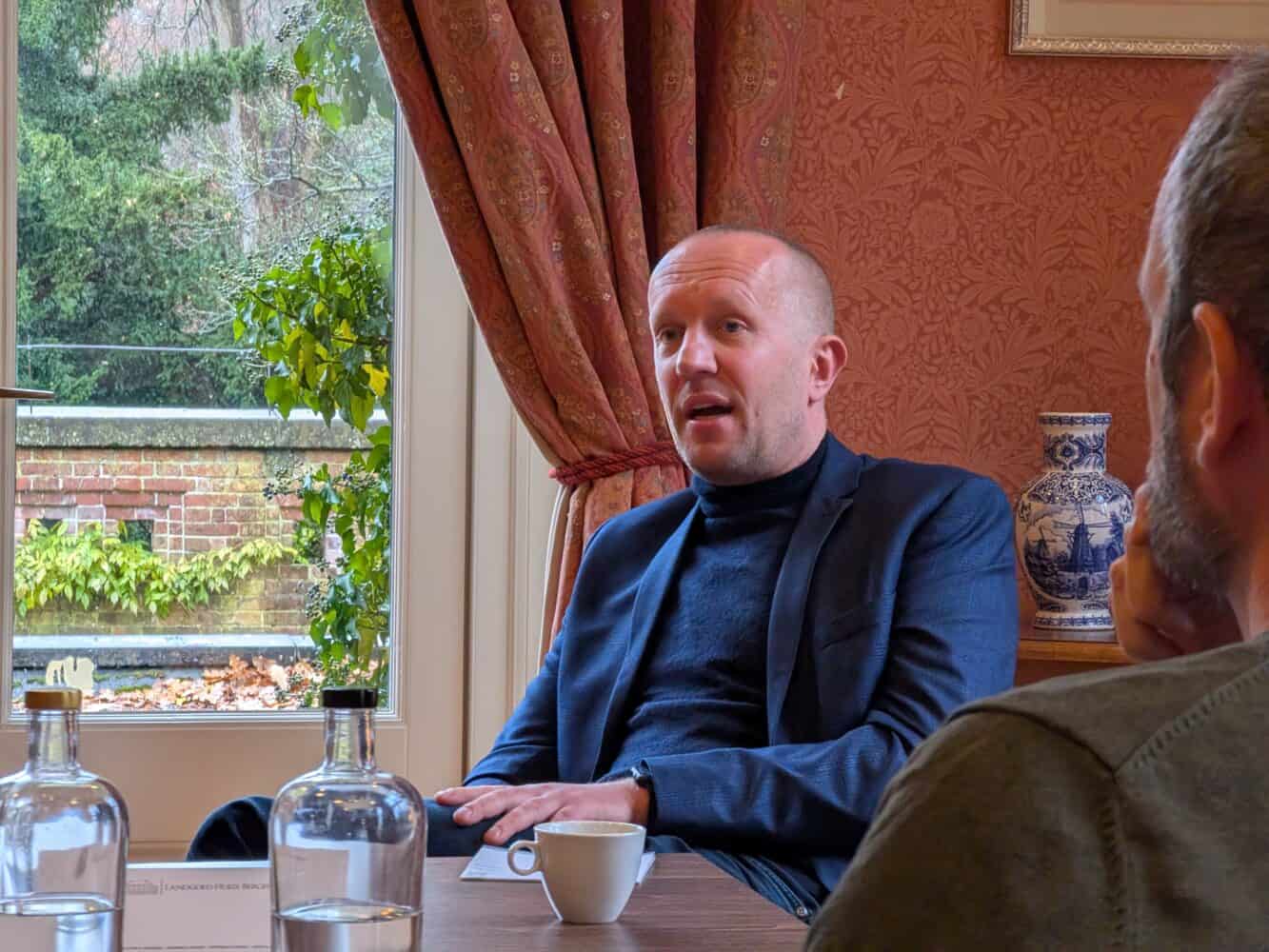 In een kamer met rode muren en een groot raam zit een man in een blauw pak aan tafel. Hij nipt van zijn koffie en bespreekt ondertussen met iemand anders strategieën voor datacenters.