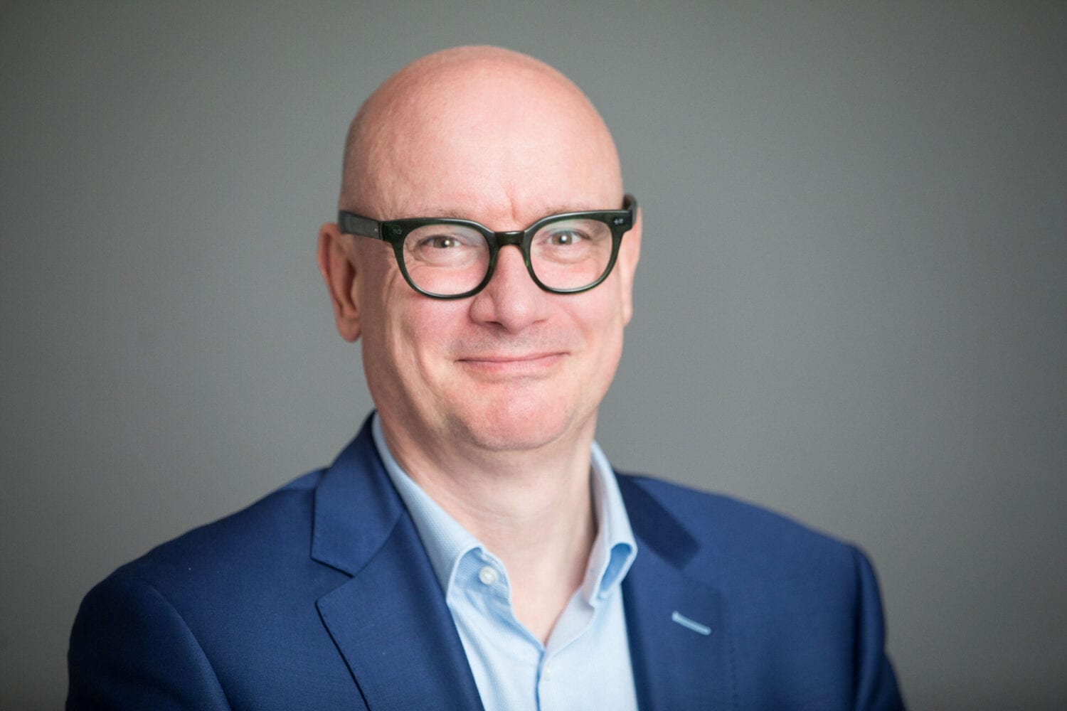 Een Cognizant-professional met een bril en een blauw colbert lacht hartelijk tegen een grijze achtergrond.