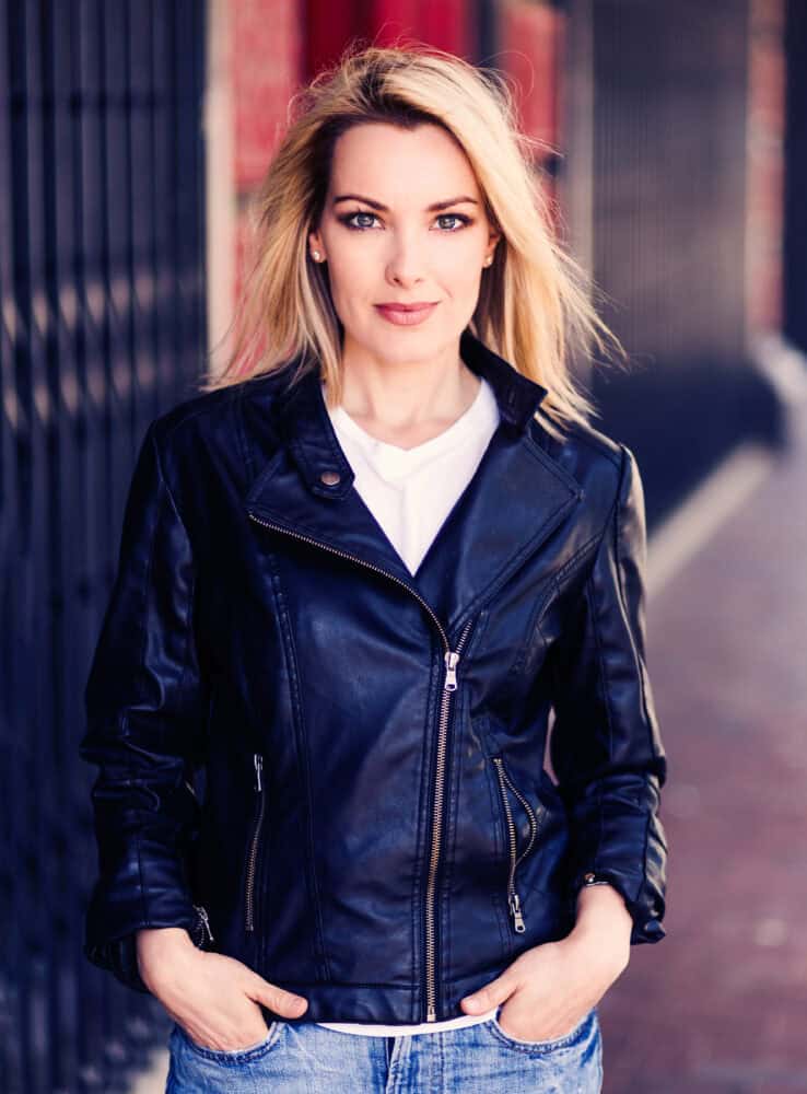 Een persoon met lang blond haar, gekleed in een zwart leren jasje en een spijkerbroek, staat zelfverzekerd buiten met zijn handen in zijn zakken, tegen een wazige stedelijke achtergrond.