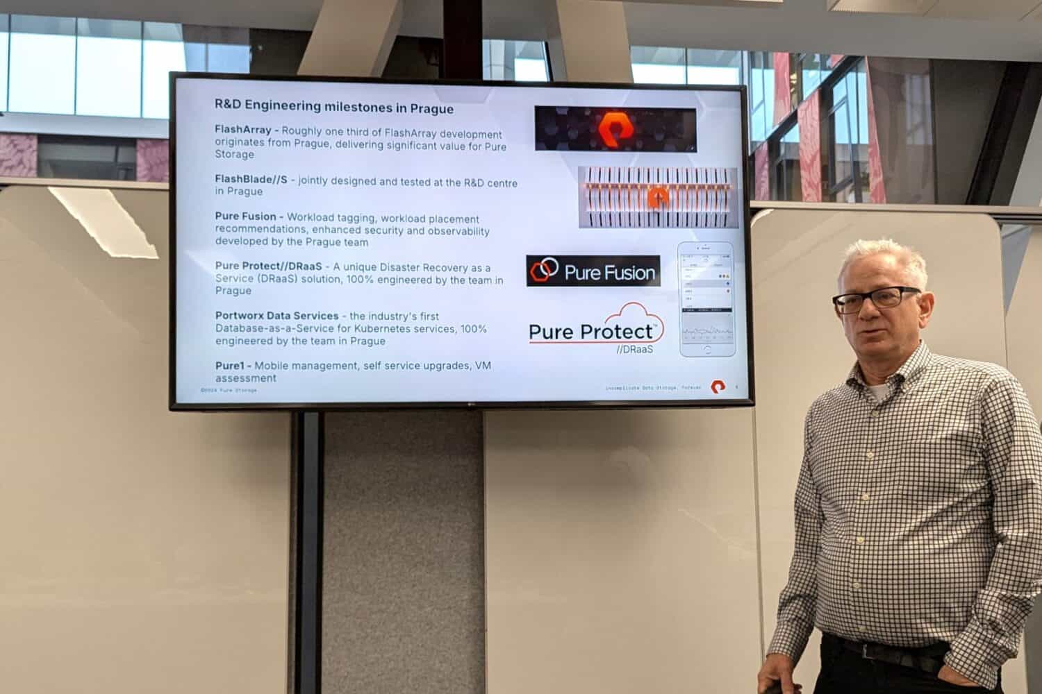 Een man staat naast een presentatiescherm met informatie over mijlpalen in de R&D-techniek in Praag, met producten als FlashArray, PureFusion en Pure Protect.