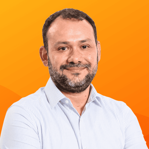 Een man met een baard, gekleed in een kenmerkend wit overhemd, staat zelfverzekerd tegen een oranje achtergrond en belichaamt de innovatieve geest van Avanade.