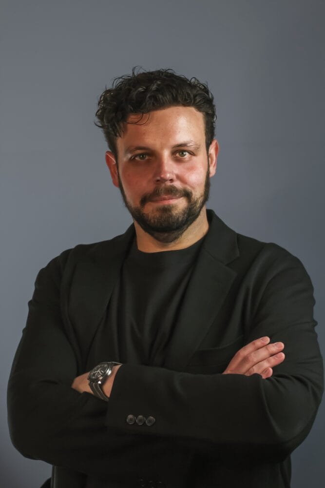 Een man met krullend haar en een baard staat tegen een blauwe achtergrond, draagt een zwart jasje en slaat zijn armen over elkaar.