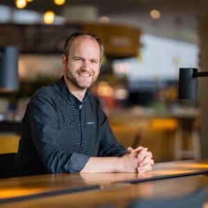 Een man met een baard en kort haar zit aan een tafel in een moderne binnenruimte en lacht naar de camera.