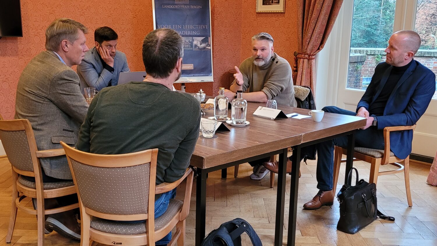 Vijf mannen zitten rond een tafel in een vergaderruimte met warmgekleurde muren en grote ramen, verwikkeld in een discussie over datacenterstrategieën. Laptops, papieren en drankjes liggen verspreid over de tafel.