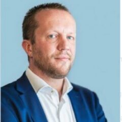 Man in een blauw colbert en wit overhemd, met gekruiste armen, tegen een lichtblauwe achtergrond.