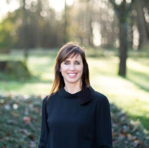Een vrouw met steil bruin haar en een zwart topje staat buiten in een zonnig park met bomen en groen op de achtergrond.