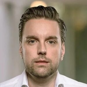 Een man met een lichte huid, kort bruin haar en lichte gezichtsbeharing kijkt recht in de camera; hij draagt een wit overhemd met kraag. De achtergrond is wazig.