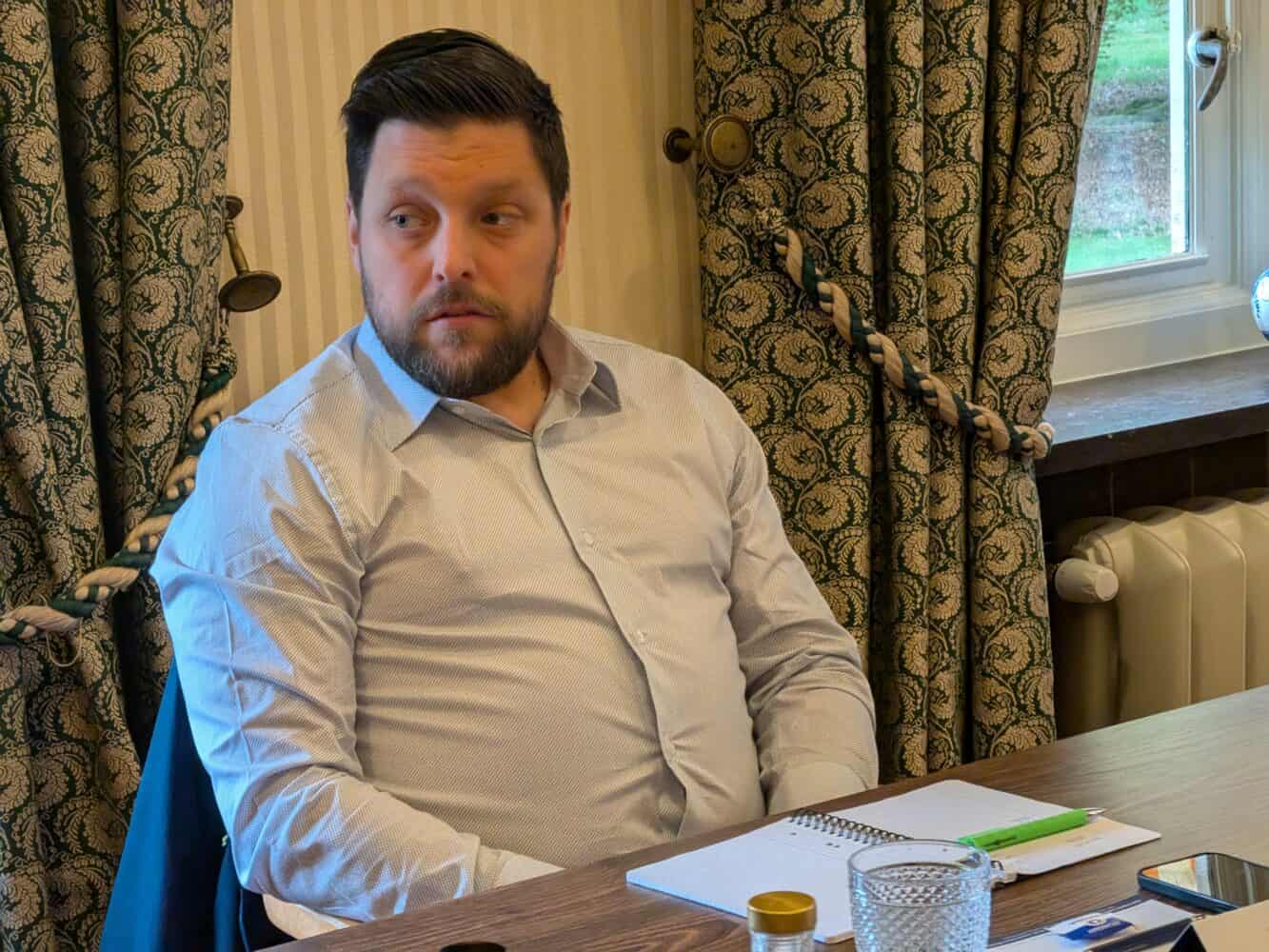 Een man met donker haar en een baard zit aan een tafel met een notitieboekje, pen en een glas water, kijkend naar links. Gordijnen met patroon en een raam zijn op de achtergrond te zien.