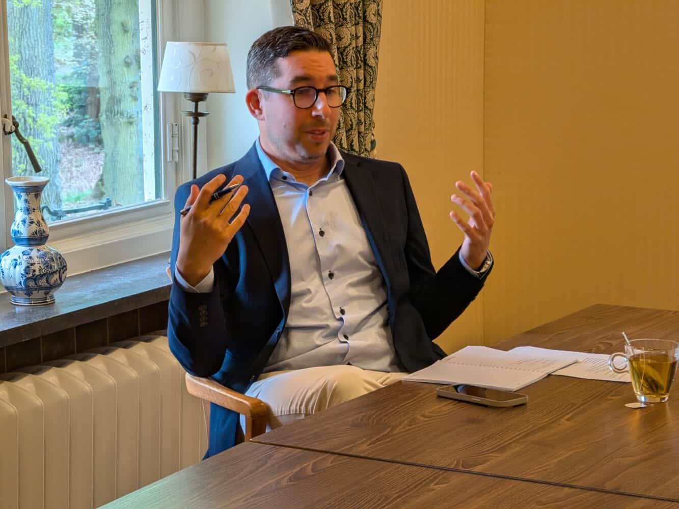 Een man in pak zit aan een tafel met papieren, een pen, een telefoon en een glas thee en maakt tijdens het spreken gebarende gebaren met beide handen.