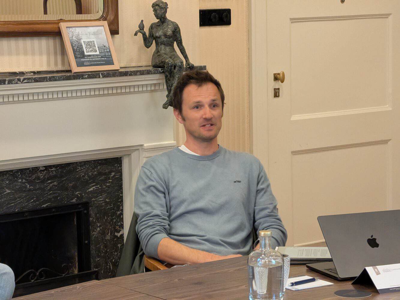 Een man in een lichtblauwe trui zit aan een tafel met een laptop, notitieboekje en waterfles, in een kamer met een open haard en een decoratief beeld.