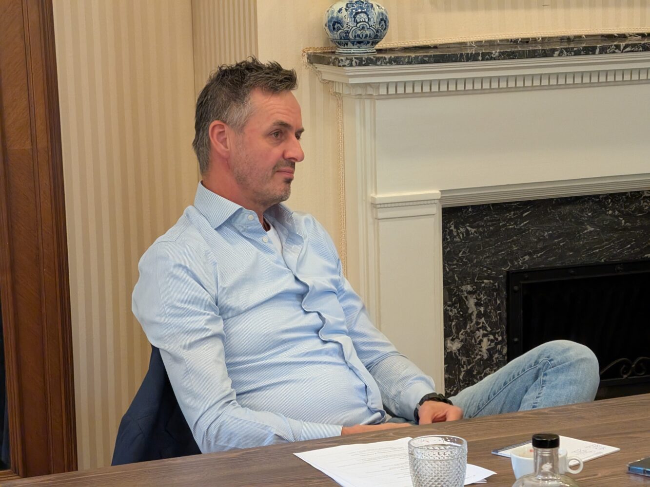 Een man in een lichtblauw shirt zit aan een tafel met papieren, een glas en een fles voor zich. Hij staat naast een open haard met een decoratieve vaas op de schouw.