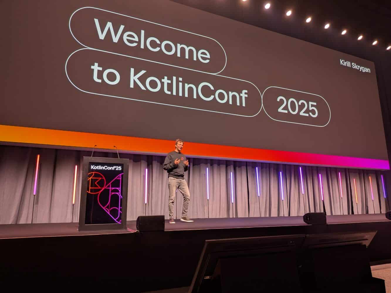 Een spreker staat op het podium voor een groot scherm met de tekst "Welcome to KotlinConf 2025", klaar om inzichten te delen over Kotlin op de conferentie.