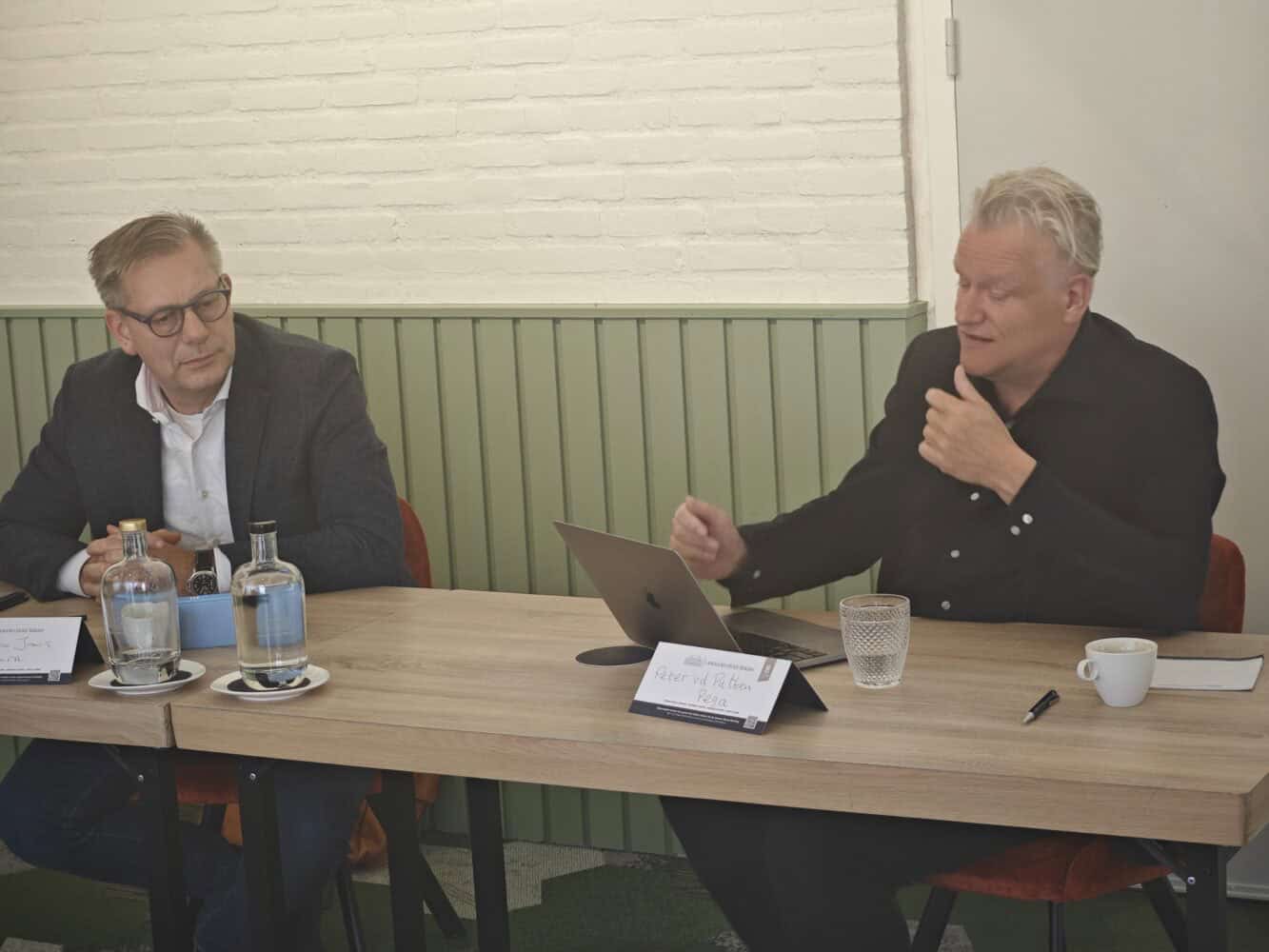 Twee mannen zitten aan een tafel in een vergaderruimte; de een spreekt en wijst naar een laptop, de ander luistert. Er liggen flessen water, een koffiekopje en papieren op tafel.
