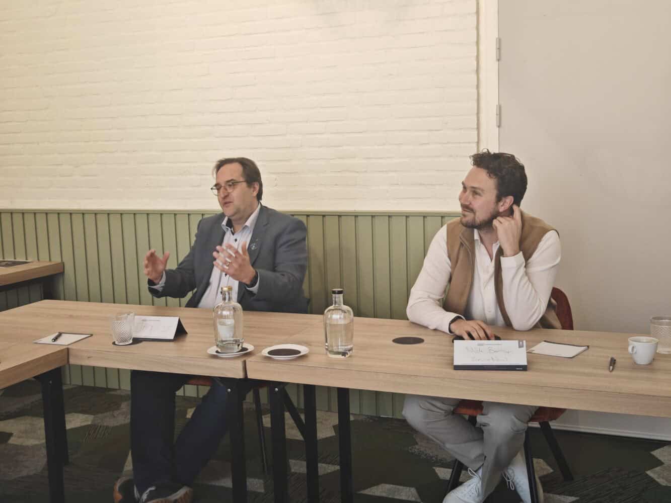 Twee mannen zitten aan een vergadertafel met notitieblokken, waterflessen en bekers voor zich. De een spreekt, de ander luistert aandachtig.