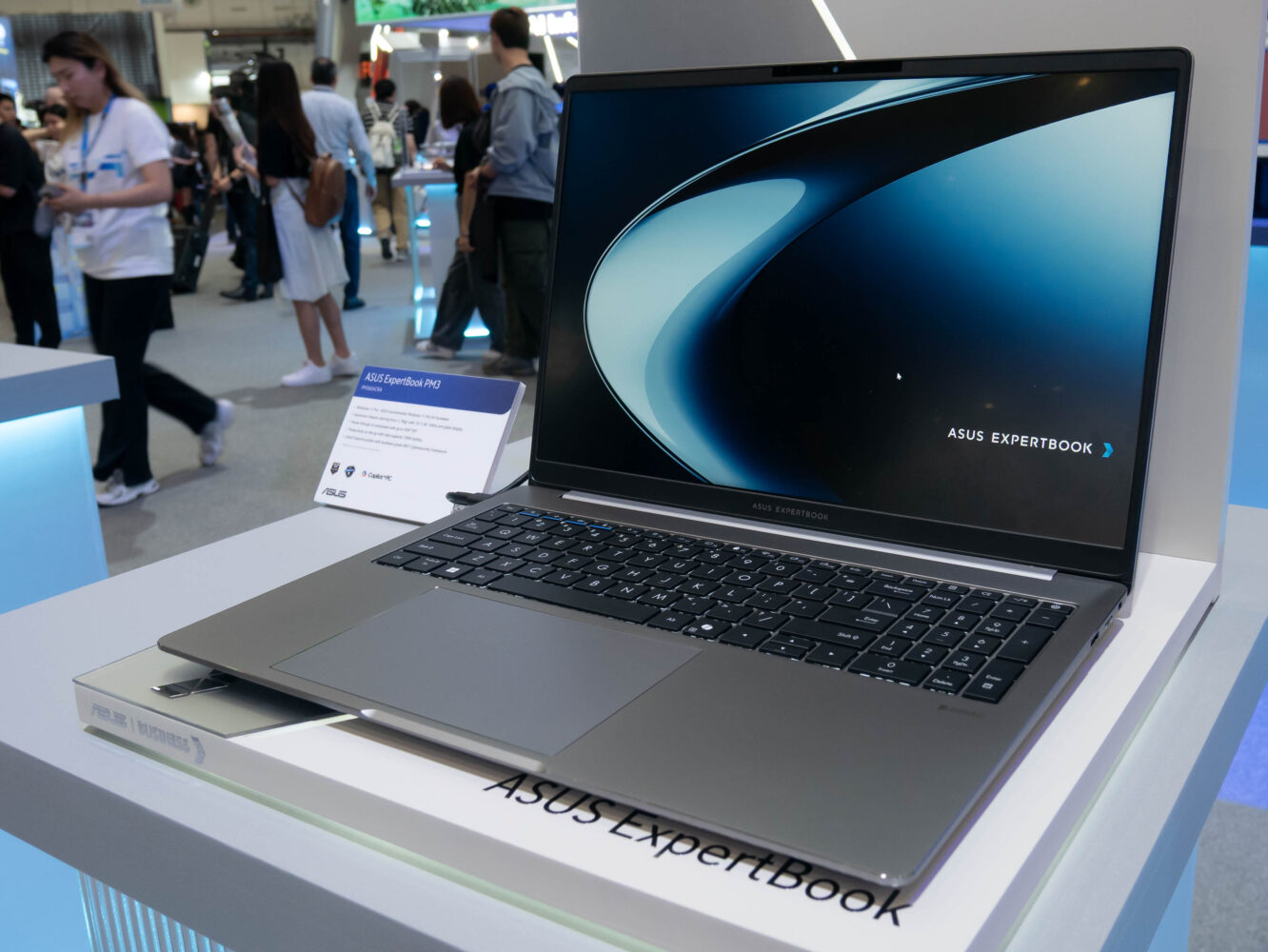 Een zilveren ASUS ExpertBook-laptop staat op een witte standaard tijdens een evenement, met het scherm aan en mensen zichtbaar op de achtergrond.