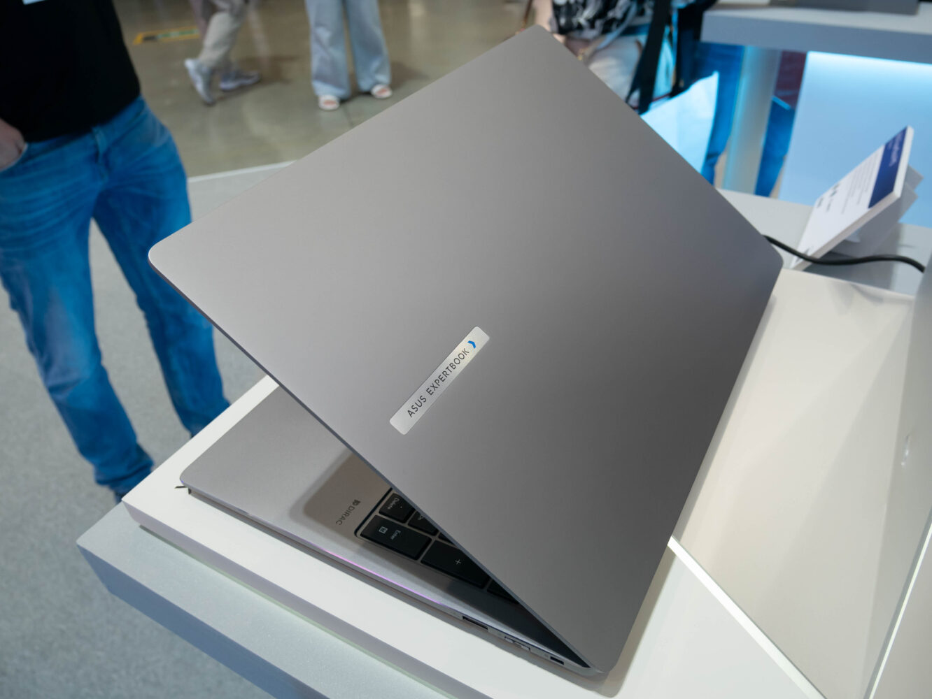 Een zilveren laptop, half opengeklapt op een presentatietafel, met een sticker met het opschrift "ASUS Zenbook". Op de achtergrond staan mensen.