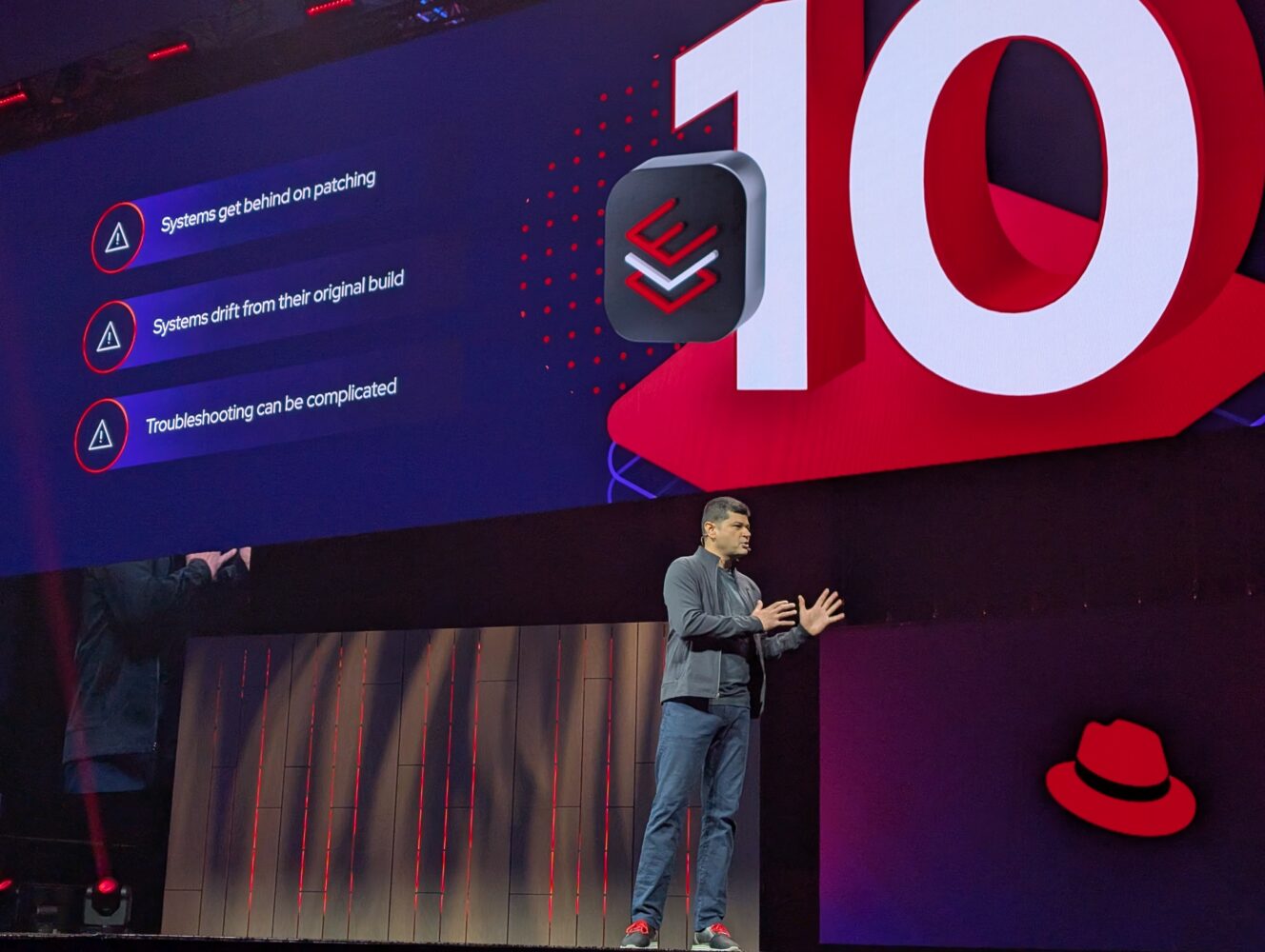 Een spreker staat op het podium voor een groot scherm met daarop het getal 10, het Red Hat-logo en een lijst met uitdagingen op het gebied van systeembeheer.