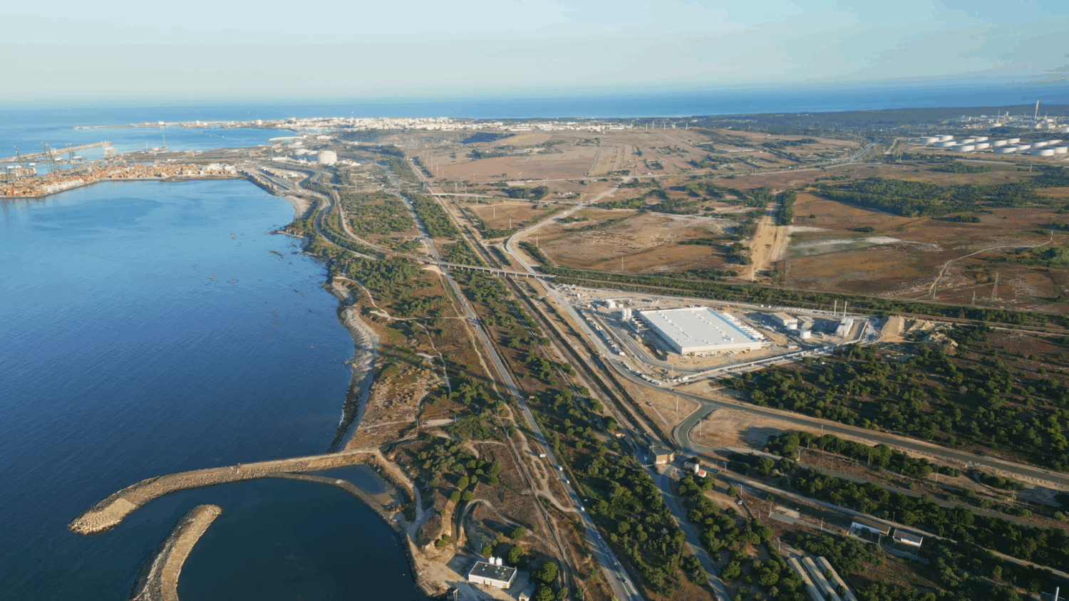 START CAMPUS 27-08 v2 4K_01_00_04_040 (Large) Luchtfoto van een industrieel gebied aan de kust met pakhuizen, wegen, groen en de zee. Aan de horizon is in de verte een stadsgezicht te zien.